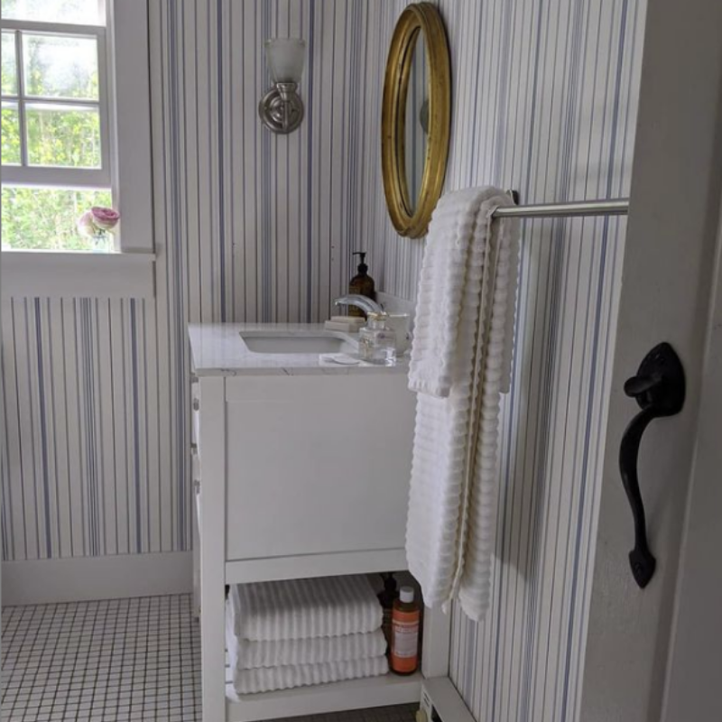 striped wallpaper in coastal bathroom with brass mirror, sconce, and window open, white towels
