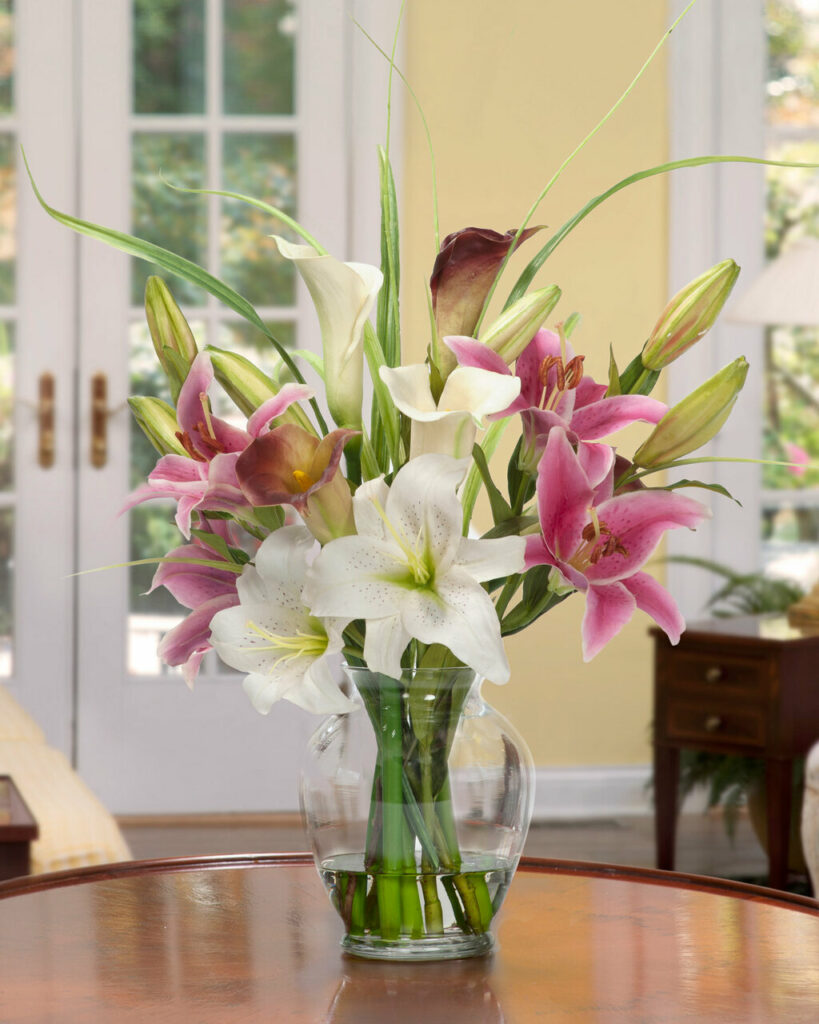 A floral arrangement in a clear vase of white and pink lilies with leaves, buds, and blooms on a brown table