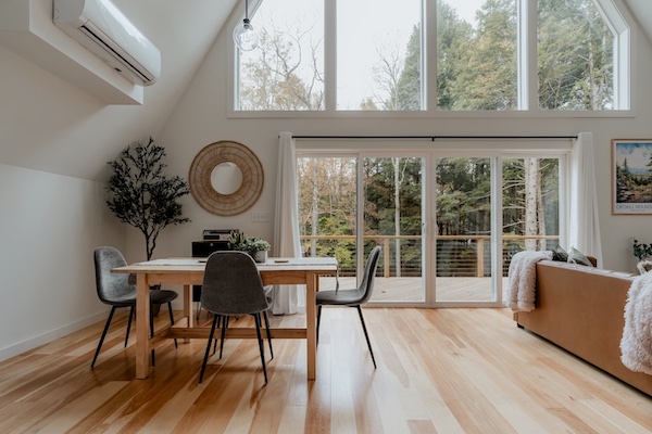 Photograph of a forest-facing dining room and living room area to demonstrate modern lake house decor, featuring Mid-Century modern dining chairs, a large plant, and a modern mirror.